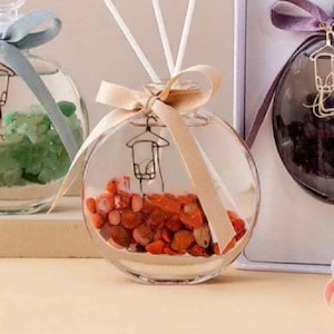 May include: Three glass diffuser bottles with decorative elements inside. The bottles are round and clear, with a ribbon tied around the neck. The first bottle contains green stones, the second bottle contains red stones, and the third bottle contains dark purple stones.