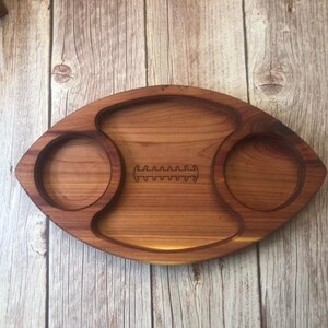 May include: A wooden serving tray shaped like a football with two circular indentations on either side. The tray is made of light brown wood and has a football seam design carved into the center.