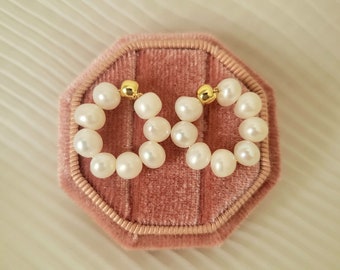 May include: A pair of pearl earrings. Each earring is composed of a circle of white pearls, connected by a small gold bead at the top. The earrings are displayed on a pink velvet jewelry box.