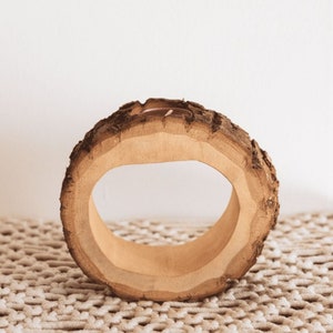 Könnte beinhalten: Ein rustikaler Serviettenring aus Holz, hergestellt aus einem Abschnitt eines Astes. Der Ring hat eine natürliche Rindenaußenseite und eine glatte, hellfarbige Innenseite. Der Serviettenring hat einen Durchmesser von etwa 10 cm und ist 4 cm dick.