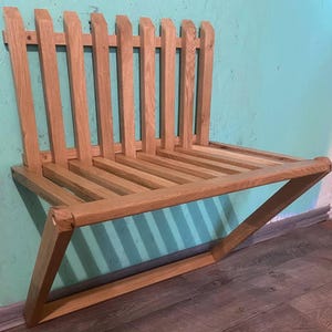 Könnte beinhalten: Eine hölzerne, an der Wand befestigte Bank mit einer Sitzfläche und Rückenlehne aus Lamellen. Die Bank ist aus hellfarbenem Holz gefertigt und hat ein einzigartiges Design mit abgewinkelten Beinen. Die Rückenlehne hat vertikale Lamellen, und die Sitzfläche besteht aus horizontalen Holzlamellen.