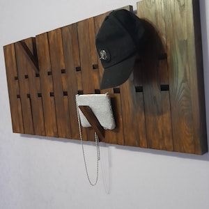 May include: A wooden wall-mounted organizer with multiple slots for hanging items. A black baseball cap hangs from one slot, a silver clutch purse hangs from another, and a silver chain hangs from a third slot.