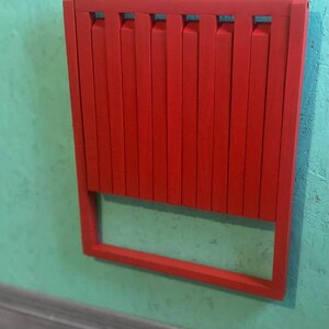 May include: A red folding chair with a slatted back and a simple frame. The chair is folded and mounted on a wall.