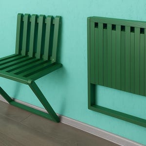 May include: A green, folding wall-mounted chair and a matching folded chair are shown against a teal wall. The chair is made of wooden slats and has a simple, modern design. The folded chair is rectangular and also made of wooden slats.