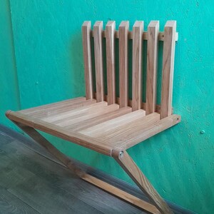 May include: A wooden wall-mounted folding bench with a slatted back and a simple design. The bench is made of light brown wood and is attached to the wall with metal brackets.