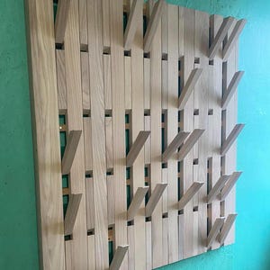 May include: A wooden wall-mounted coat rack with a unique design. The rack is made of light-colored wood and features a series of vertical slats with angled hooks that fold out. The hooks are arranged in a staggered pattern, creating a visually interesting and functional design.
