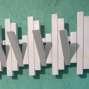 Può includere: Un appendiabiti in legno bianco con un design unico che presenta più listelli verticali con ganci angolati. L'appendiabiti è montato su una parete verde.