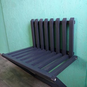 May include: A dark gray wooden wall-mounted shelf with a fold-down seat. The shelf has a slatted design and is attached to the wall with screws.