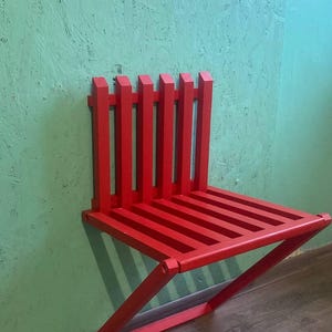 May include: A bright red, wooden, folding wall-mounted chair. The chair is made of slats and has a simple, modern design.