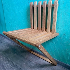 May include: A wooden wall-mounted folding chair with a slatted back and a triangular base. The chair is made of light brown wood and is mounted to a turquoise wall.