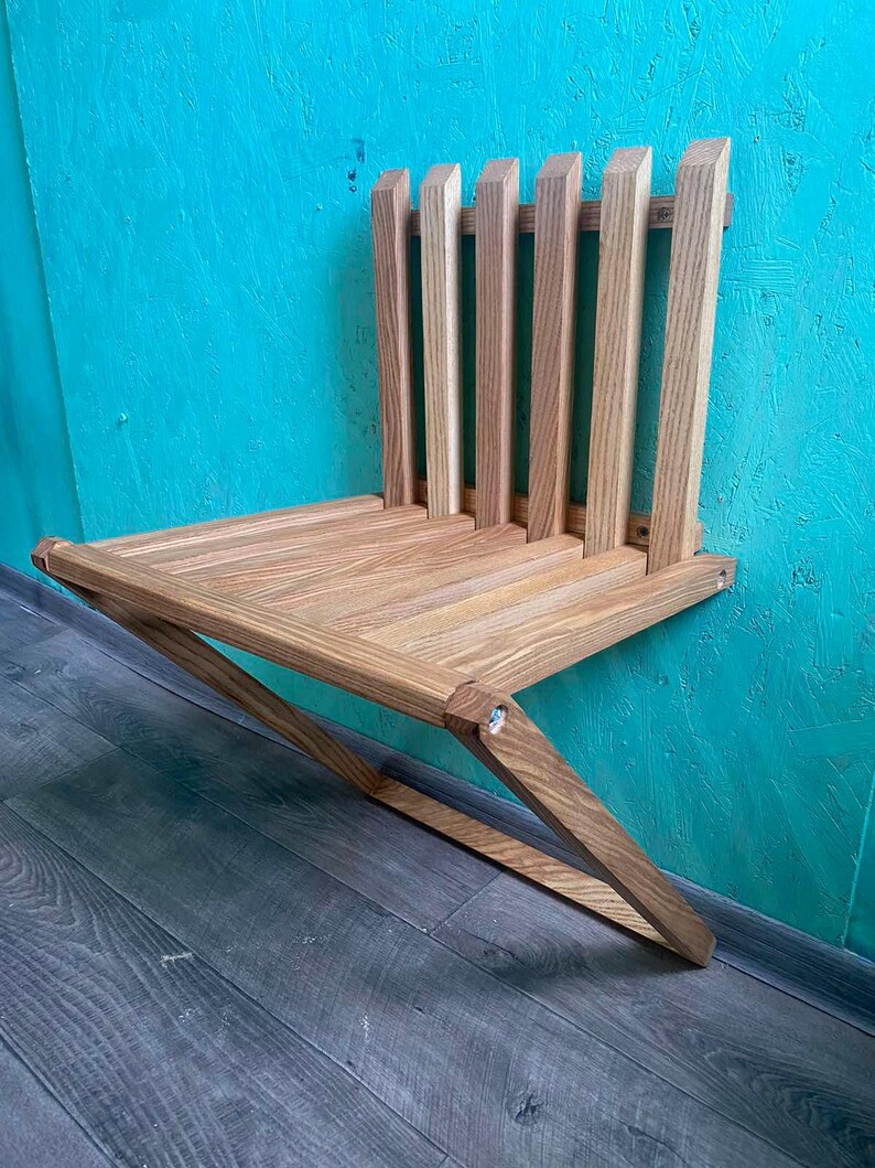 May include: A wooden wall-mounted folding chair with a slatted back and a triangular base. The chair is made of light brown wood and is mounted to a turquoise wall.