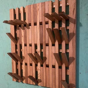 以下が含まれることがあります： ユニークなデザインの壁掛け式木製コートラック。ラックは、グリッドパターンに配置された複数の木製の羽根板で構成されています。羽根板は濃い茶色に染色されており、自然な木目があります。ラックには、外側に角度が付けられた複数のフックがあり、コート、帽子、バッグを掛けるのに十分なスペースがあります。