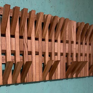 Può includere: Un attaccapanni da parete in legno dal design unico. L'attaccapanni è realizzato in legno marrone chiaro e presenta più ganci angolati. Il design è moderno e geometrico, con una serie di elementi in legno verticali e angolati.