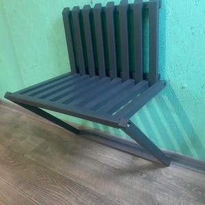 May include: A dark gray metal wall-mounted folding bench with a slatted design. The bench is folded up and mounted to a wall.