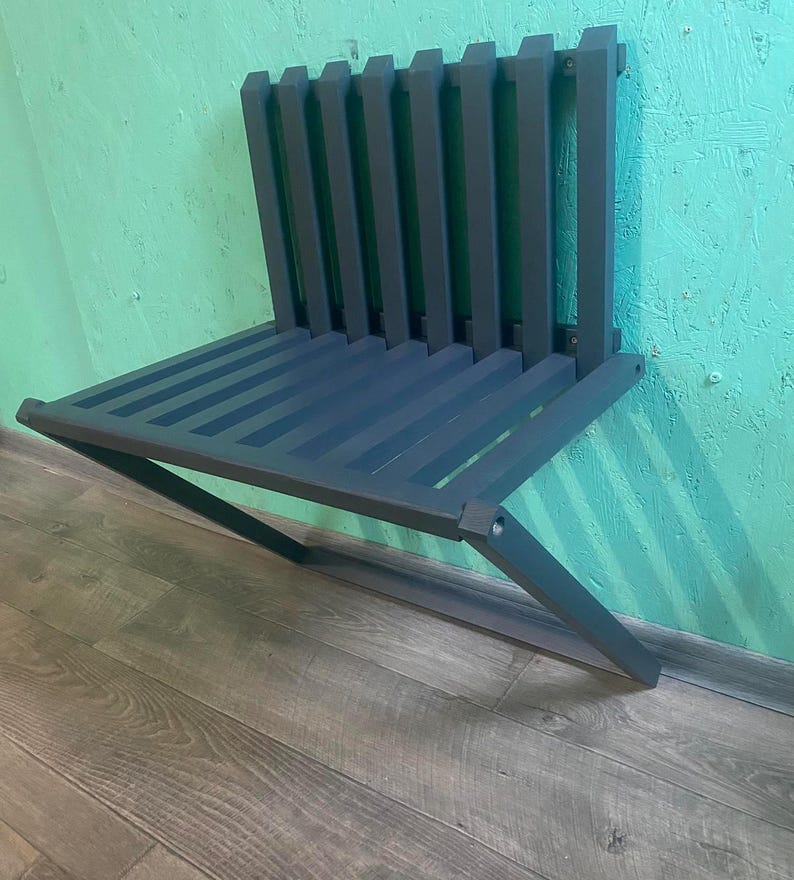 May include: A dark gray metal wall-mounted folding bench with a slatted design. The bench is folded up and mounted to a wall.