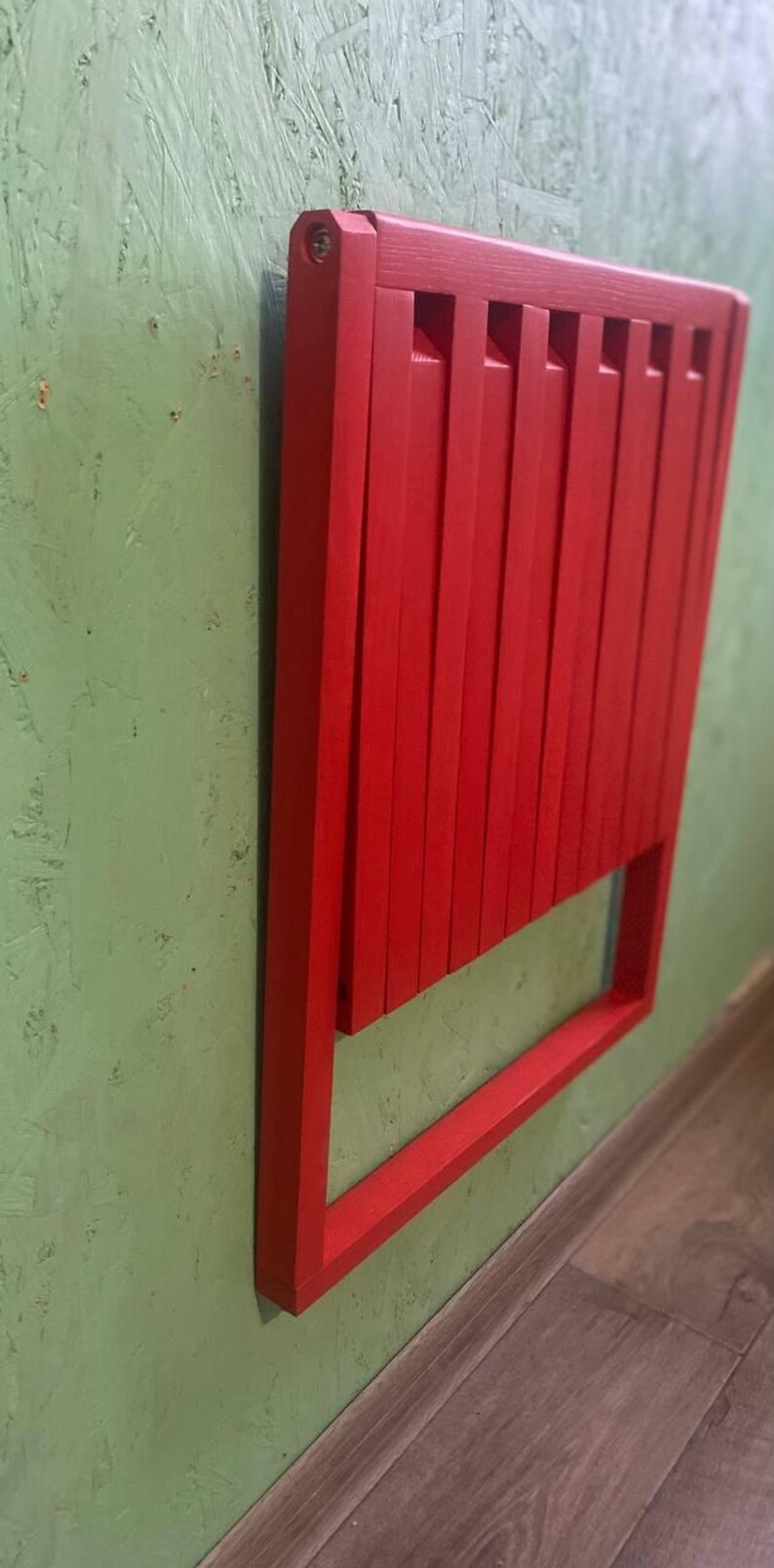 May include: A red wooden folding chair is mounted to a green wall. The chair is folded and has a simple design.