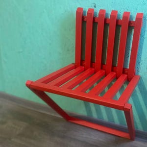May include: A red folding chair with a slatted back and seat. The chair is folded and leaning against a wall.