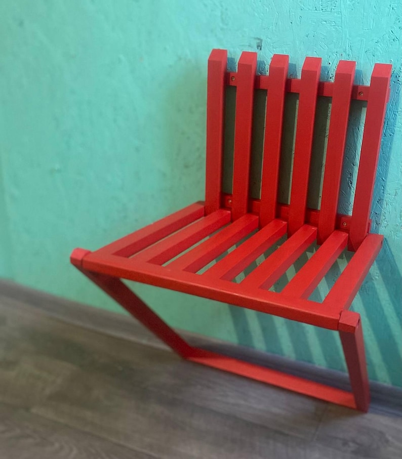 May include: A red folding chair with a slatted back and seat. The chair is folded and leaning against a wall.