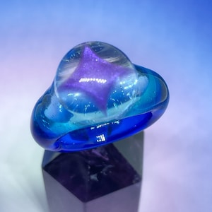Puede incluir: Un anillo de vidrio azul con una cúpula transparente que contiene una estrella morada. El anillo está sobre un soporte morado.