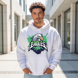 Könnte beinhalten: Ein weißer Kapuzenpullover mit einem Grafikdruck eines Adlerkopfes mit gelbem Schnabel und grünen sowie schwarzen Farbspritzern. Das Wort EAGLES ist in einer fetten, stilisierten Schriftart in der Mitte des Adlermotivs aufgedruckt.