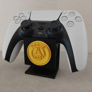 Peut inclure: Une manette PlayStation 5 blanche et noire posée sur un support noir avec un logo Paris Saint-Germain doré. La manette a des joysticks et des boutons noirs. Le support est rectangulaire et maintient la manette à la verticale.