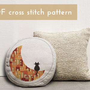 May include: A round cushion featuring a cross stitch design of a black cat sitting on a crescent moon made of orange and yellow houses. The cushion is on a white surface next to a beige cushion.
