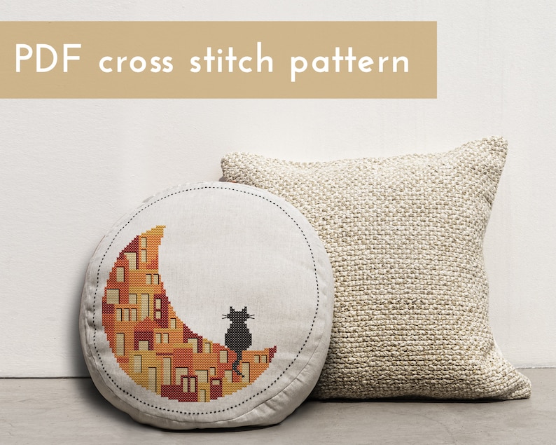 May include: A round cushion featuring a cross stitch design of a black cat sitting on a crescent moon made of orange and yellow houses. The cushion is on a white surface next to a beige cushion.