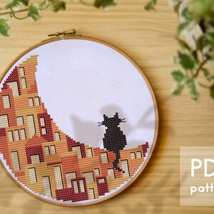 May include: A cross-stitch pattern featuring a black cat sitting on a crescent moon with a geometric design in shades of orange, yellow, and red.