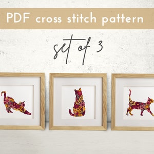 May include: Set of three cross stitch patterns featuring floral cat silhouettes in shades of red, yellow, and orange. The patterns are framed and displayed on a white wall.
