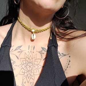 Könnte beinhalten: Handgefertigte Makramee-Halskette in Hellgrün mit einem kleinen weißen Muschelanhänger. Die Halskette hat ein zartes Webmuster und ist mit kleinen goldfarbenen Perlen verziert. Der Hintergrund ist ein schwarzes Oberteil und ein Blumen-Tattoo.