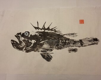 Puede incluir: Una impresión de pez en tinta negra sobre papel blanco roto, que muestra un pez detallado con aletas dorsales espinosas, boca abierta y un ojo visible. Un pequeño sello rojo cuadrado se encuentra en la esquina superior derecha del papel.