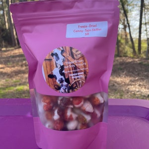 May include: A pink, resealable bag of freeze-dried Camoy-Tajin Skittles. The bag has a clear window showing the colorful contents. A circular label features a cow illustration and the text "Three Crazy Heifers Treats".