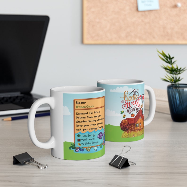 May include: Two white ceramic mugs with different designs. The mug on the left has a pixelated design of a video game character with the text "Water Artisan Goods Essential for life is Pelican Town and your Stardew Valley adventure Keep your crops growing and your energy flowing +200 Energy +105 Health +30 Max Energy". The mug on the right has a cartoon illustration of a house with the text "Home Sweet Home".