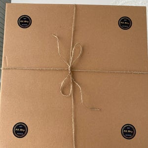 May include: A rectangular, brown cardboard box tied with twine. Four black and white circular stickers with the text "ART M.A. Art Design" are placed on the corners of the box. The twine is tied in a bow.