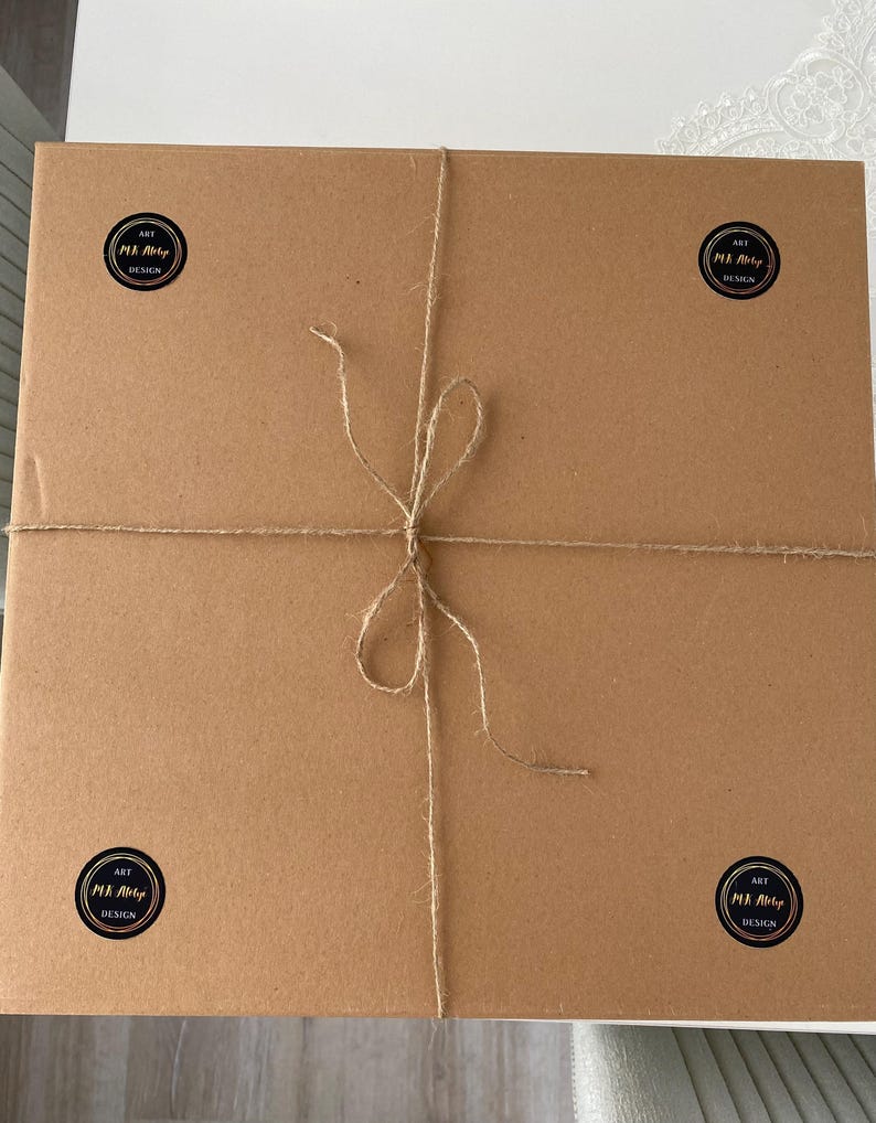 May include: A rectangular, brown cardboard box tied with twine. Four black and white circular stickers with the text "ART M.A. Art Design" are placed on the corners of the box. The twine is tied in a bow.