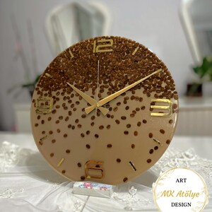 May include: Round wall clock with a coffee bean design. The clock face is a gradient of brown and tan, with gold-colored numerals and hands. The clock is decorated with whole coffee beans, creating a unique and textured appearance. The clock is a decorative item.