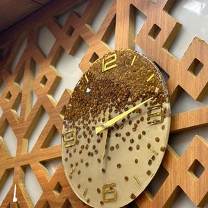May include: A round wall clock with a clear face, filled with coffee beans. The clock has gold-colored hands and numerals. It is mounted on a wooden wall with a geometric pattern.