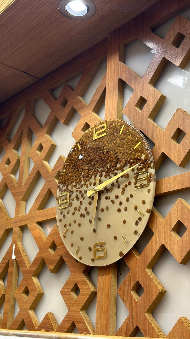 May include: A round wall clock with a clear face, filled with coffee beans. The clock has gold-colored hands and numerals. It is mounted on a wooden wall with a geometric pattern.