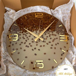 May include: A round wall clock with a coffee bean design. The top half of the clock face is filled with coffee beans, while the bottom half is a light tan color. Gold-colored hands and numerals mark the time. The clock is in a cardboard box.