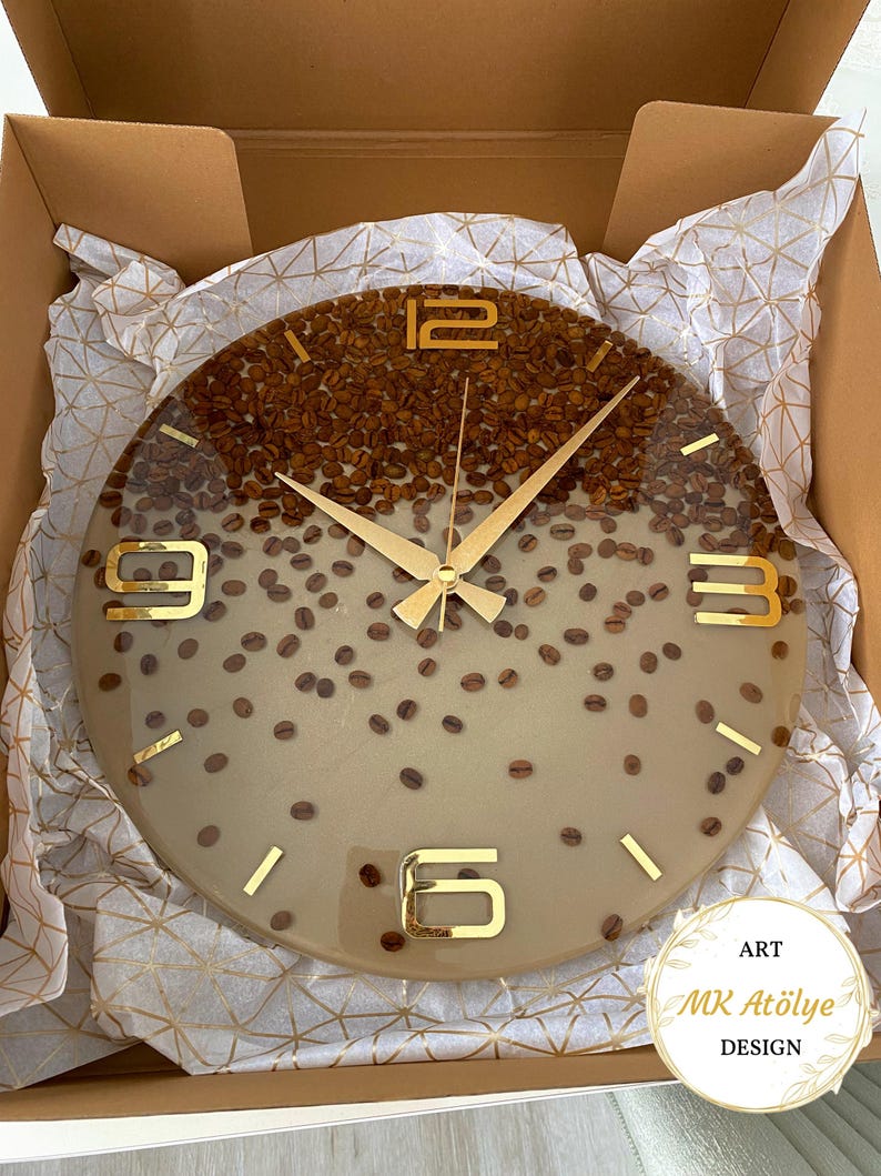 May include: A round wall clock with a coffee bean design. The top half of the clock face is filled with coffee beans, while the bottom half is a light tan color. Gold-colored hands and numerals mark the time. The clock is in a cardboard box.