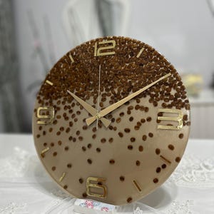 May include: A round wall clock with a coffee bean design. The clock face is a gradient of tan and brown, with embedded coffee beans. Gold-colored numerals and hands complete the design. The clock is a decorative item.