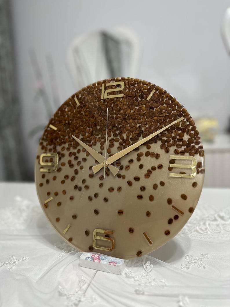 May include: A round wall clock with a coffee bean design. The clock face is a gradient of tan and brown, with embedded coffee beans. Gold-colored numerals and hands complete the design. The clock is a decorative item.