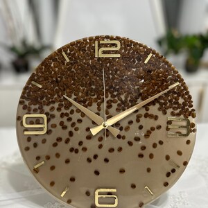 May include: A round wall clock with a coffee bean design. The clock face is a gradient of brown and tan, embedded with coffee beans. Gold-colored hands and numerals mark the time. The clock is a decorative item.