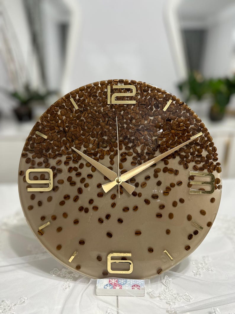 May include: A round wall clock with a coffee bean design. The clock face is a gradient of brown and tan, embedded with coffee beans. Gold-colored hands and numerals mark the time. The clock is a decorative item.
