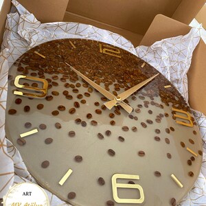May include: A round wall clock with a coffee bean design. The clock face is a light beige color with embedded coffee beans and gold-colored numerals and hands. The clock is packaged in a cardboard box with decorative paper.