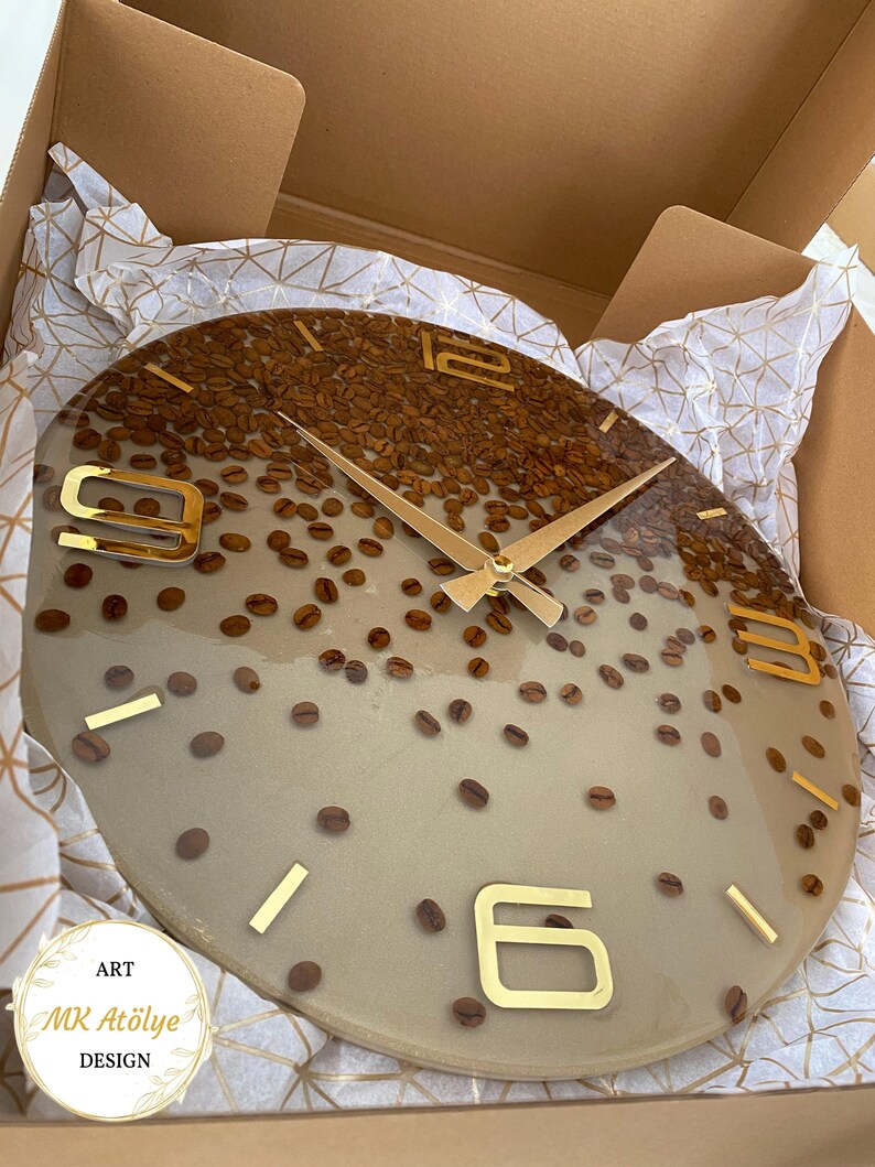 May include: A round wall clock with a coffee bean design. The clock face is a light beige color with embedded coffee beans and gold-colored numerals and hands. The clock is packaged in a cardboard box with decorative paper.