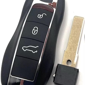 Puede incluir: Llave de coche negra con ribete plateado y tres botones: desbloquear, bloquear y abrir el maletero. Una hoja de llave de metal separada está al lado del llavero. La llave tiene un diseño elegante y moderno.