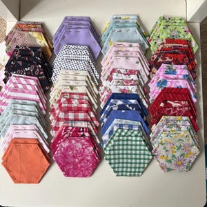 May include: A collection of fabric hexagons arranged in stacks, featuring a variety of colours and patterns including solid orange, pink batik, green gingham, blue plaid, floral prints, polka dots, stripes, and paisley designs, all basted with thread.
