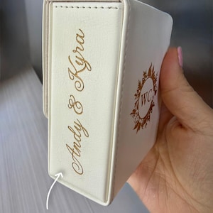 May include: Ivory-colored, rectangular box with gold lettering that reads "Andy & Kyra" on the side. The other side features a gold floral design with the initials "WC" inside. The box appears to be made of leather or a similar material.