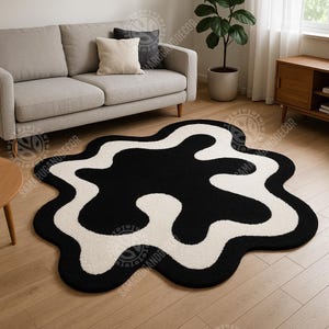 May include: A black and white abstract area rug with a unique, organic shape. The rug features a bold black design against a cream-colored background. The rug is placed on a light-colored hardwood floor, complementing the neutral tones of the room's decor.